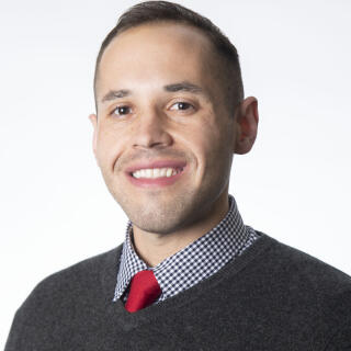 Man wearing a checkered shirt and red tie under a black sweater in front of a white background