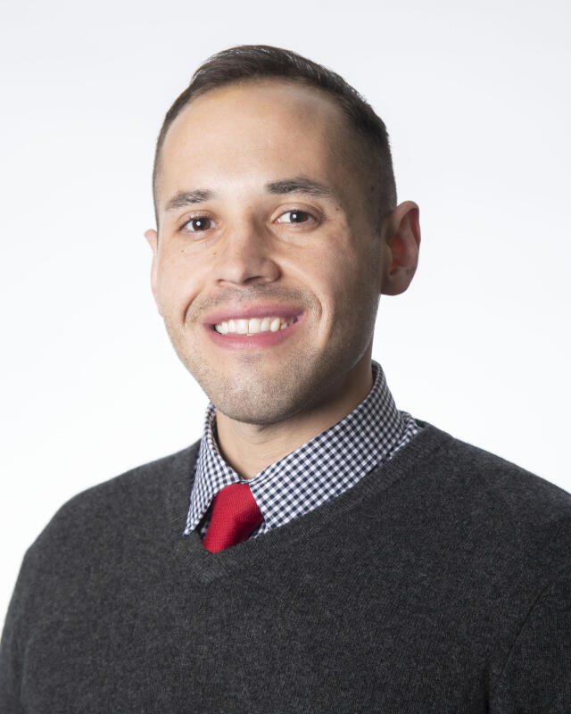 Man wearing a checkered shirt and red tie under a black sweater in front of a white background