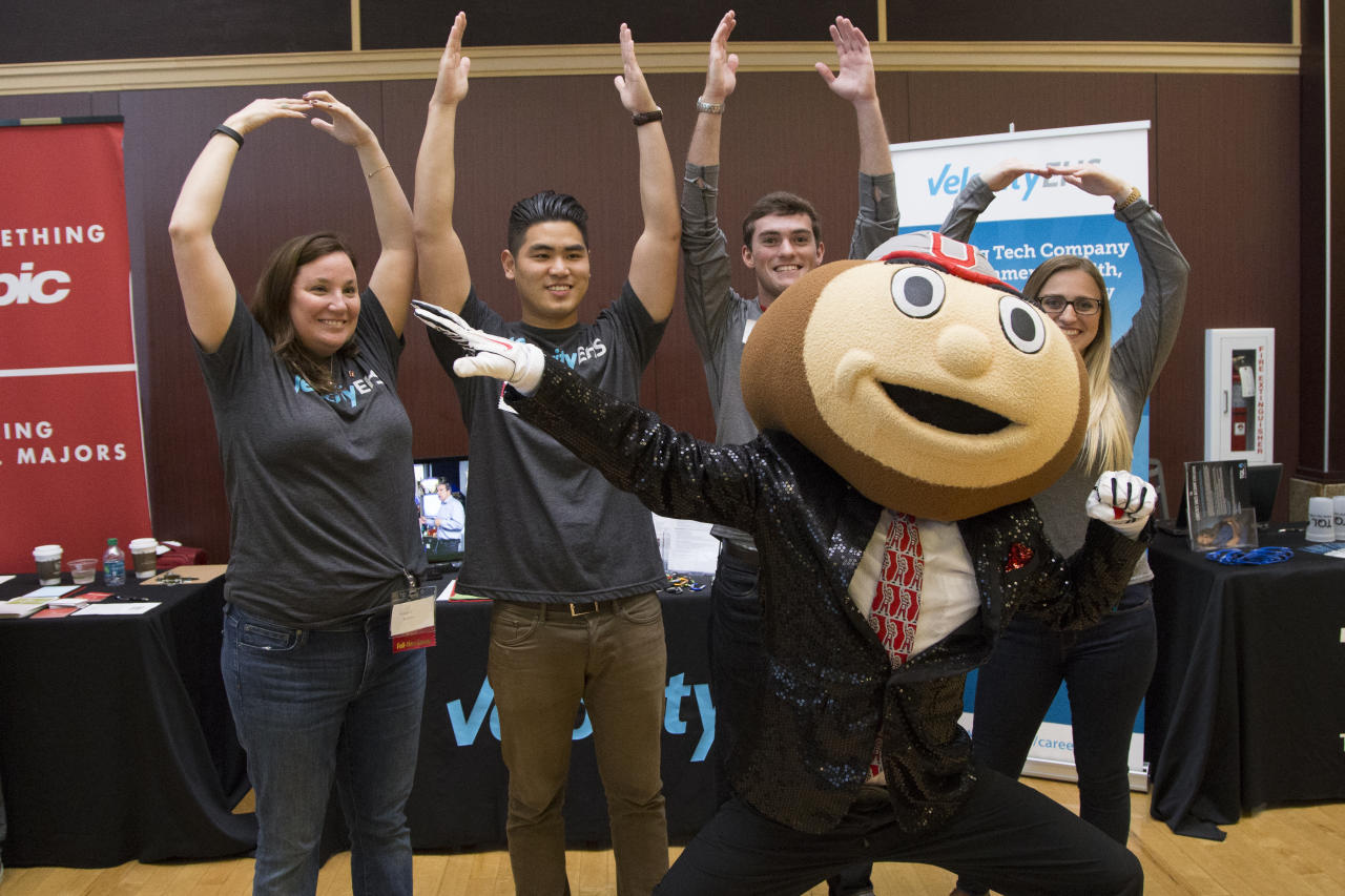 Brutus Buckeye poses for a photo in front of four employers at a career fair on campus